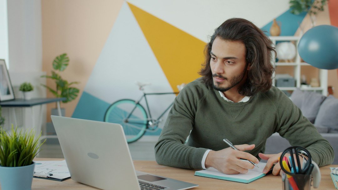 Remote worker taking notes in a notebook beside a laptop